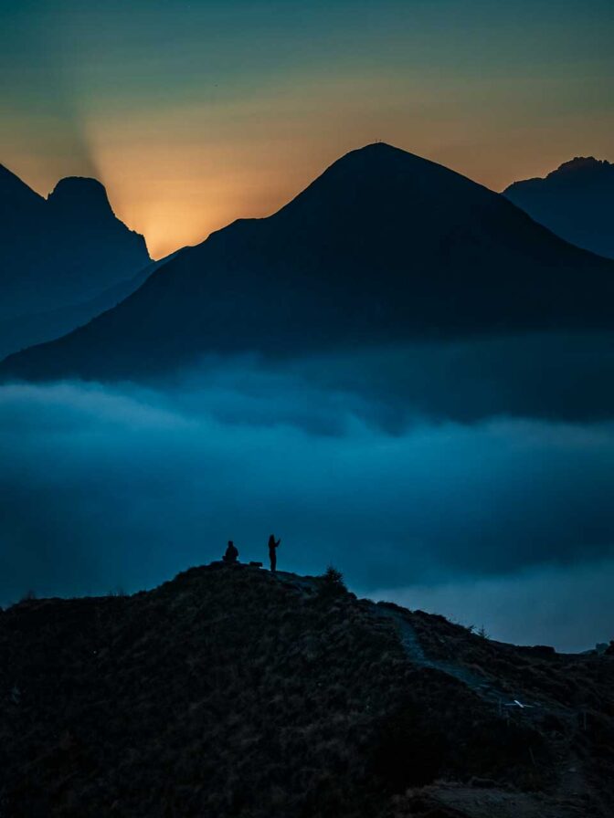 Die Sonne ist hinter der Marmolata untergegangen. Vor blauem Nebel hocken noch einige Fotografen. Am Gipfel des Monte Pore ist das Gipfelkreuz zu erkennen.